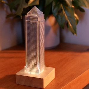Puede incluir: Una maqueta transparente de rascacielos en miniatura con una fachada texturizada, iluminada desde dentro, se asienta sobre una base cuadrada de madera. El edificio tiene una parte superior puntiaguda. El fondo presenta una planta verde.