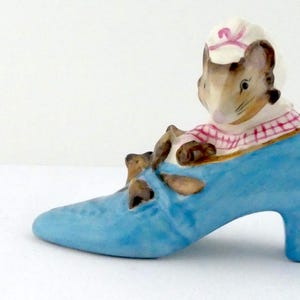 May include: A ceramic mouse figurine in a light blue high-heeled shoe. The mouse wears a pink and white checkered shirt and a white hat with a pink ribbon. The shoe has brown details and a small heel.