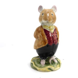 Peut inclure: Figurine en céramique représentant une souris vêtue d'une veste marron, d'un gilet rouge et d'un pantalon noir. La souris a un visage brun clair et des oreilles roses. Elle se tient sur une base verte et jaune.