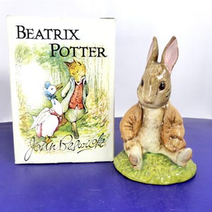 May include: A ceramic figurine of a brown rabbit wearing a tan jacket, sitting on a green base, accompanied by a rectangular box featuring the text Beatrix Potter and John Beswick with an illustration of a duck and a fox in a forest setting.