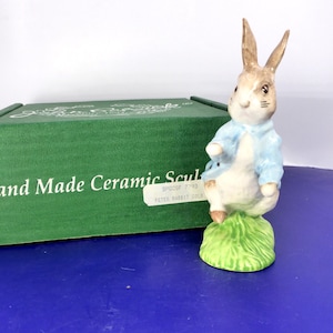 May include: A ceramic figurine of a rabbit wearing a light blue jacket, standing on a green grassy base. Behind the figurine is a green box with the text Hand Made Ceramic Sculpture and a label reading BPGSF 7793 PETER RABBIT GOLD.