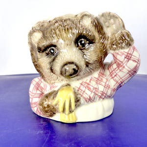 May include: A ceramic figurine of a brown bear cub with a glossy finish. The bear is wearing a white shirt with a red plaid pattern and has one paw raised near its head. The bear has dark eyes, a black nose, and a yellow scarf around its neck.