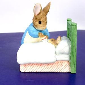 Peut inclure: Une figurine en céramique représentant un lapin brun portant une veste bleue debout à côté d'un lit. Un plus petit lapin brun est couché dans le lit sous une couverture blanche. Le lit a une tête de lit verte et une base à rayures orange.