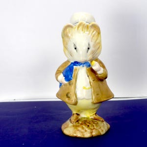 Peut inclure: Une figurine en céramique représentant une souris vêtue d'un manteau beige, d'un gilet blanc et d'un foulard bleu. La souris a un pelage brun clair, des oreilles pointues et se tient sur une base texturée. Elle porte un chapeau blanc.