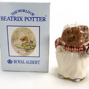 May include: A ceramic figurine of a hedgehog wearing a red and white plaid shirt and a white bonnet, holding a gray object. The figurine is part of the "World of Beatrix Potter" collection by Royal Albert. A matching box is to the left.