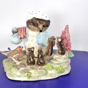 May include: A ceramic figurine featuring a hedgehog wearing a white bonnet, blue jacket, and patterned apron, holding a brown bag. The hedgehog is surrounded by a robin, two small brown mice, and a larger mouse near a pink floral plant.
