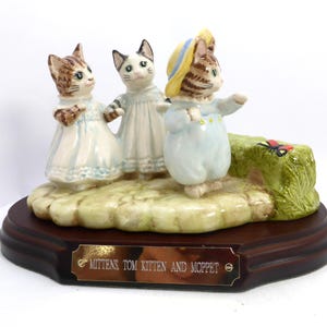 May include: A ceramic figurine featuring three anthropomorphic kittens on a base. The kittens are dressed in pastel clothing and stand on a grassy area. A plaque on the base reads "Mittens Tom Kitten and Moppet."