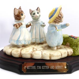 May include: A ceramic figurine featuring three anthropomorphic kittens. The kittens are dressed in light blue and white outfits, standing on a textured base. A butterfly rests on a green patch. The base is dark brown with a gold plaque that reads "MITTENS TOM-KITTEN AND MOPPET."