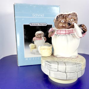 May include: A ceramic Schmid Beatrix Potter musical figurine of Mrs. Tiggy-Winkle the hedgehog wearing a white apron and pink checkered shawl, standing on a stone-patterned base next to a small basket, accompanied by its original blue cardboard box.
