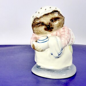 May include: A ceramic hedgehog figurine wearing a white bonnet with brown spots, a pink and white checked shirt, and a white apron. The hedgehog is holding a small white teapot with blue trim and a white cloth in its paws, standing on a base.
