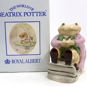 May include: A ceramic figurine of a frog wearing a pink coat and green vest, seated on a stone bench. The figurine is from the "World of Beatrix Potter" collection by Royal Albert. The box features an illustration of a frog.