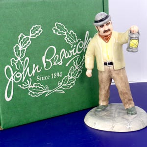 May include: A ceramic figurine of a man with a mustache wearing a light yellow jacket, brown trousers, and a gray hat, holding a small lantern with a yellow light. The figurine stands on a gray cobblestone base next to a green John Beswick box.