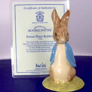 Op de afbeelding: Een keramieken beeldje van Pieter Konijn in een blauw jasje, staand op een groene ondergrond. Achter het beeldje staat een certificaat van echtheid van Beswick, The World of Beatrix Potter, getiteld Sweet Peter Rabbit, met tekst en logo's.