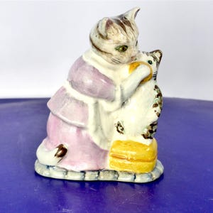 May include: A ceramic figurine depicting a larger cat wearing a light purple garment, standing and holding a smaller white and brown tabby kitten. The kitten is positioned on a yellow rectangular base. The cats have painted details.