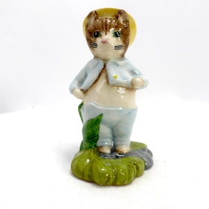 Può includere: Statuina in ceramica di un gatto con cappello giallo e abito azzurro. Il gatto ha pelo marrone e bianco e occhi verdi. È in piedi su una base verde e grigia. Un oggetto decorativo.