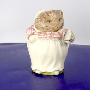 May include: A ceramic figurine of a brown hedgehog wearing a white bonnet and a pink and white checked shirt over a long white apron. The hedgehog is holding a small white cup in one hand, featuring detailed facial features and textured fur.