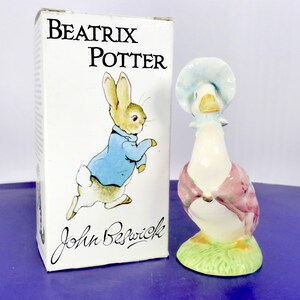 Puede incluir: Una caja de cartón blanca con el texto Beatrix Potter y John Beswick y una ilustración de un conejo con chaqueta azul, junto a una figura de cerámica de un ganso blanco con un gorro azul y un chal rosa sobre una base verde.