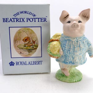 Puede incluir: Una figura de cerdo de cerámica con una chaqueta azul texturizada y una cesta amarilla, sobre una base verde. Al lado hay una caja azul claro con el texto The World of Beatrix Potter y Royal Albert, que incluye una ilustración de una rana.