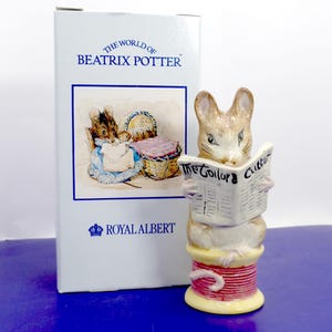Puede incluir: Una figura de ratón de cerámica sentada sobre un carrete de hilo rojo y amarillo, sosteniendo un periódico blanco con el texto The Tailor & Cutter. Al lado hay una caja rectangular blanca con la etiqueta The World of Beatrix Potter y Royal Albert.