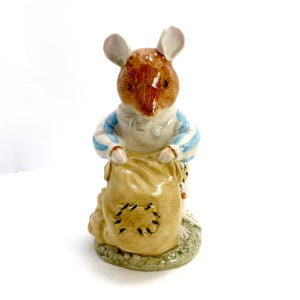 May include: A ceramic figurine of a mouse wearing a blue and white striped shirt, holding a tan sack with black stitched patches. The mouse has brown fur and pink ears, standing on a green base.