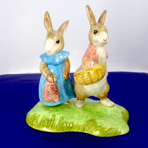 May include: A ceramic figurine featuring two rabbits standing on a green grassy base. The left rabbit wears a blue dress and carries a small pink bag. The right rabbit wears a pink jacket and carries a yellow woven basket. Both have brown fur.