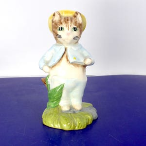 May include: A ceramic figurine of a brown tabby cat standing upright on a green grassy base. The cat wears a yellow hat, a light blue jacket, and light blue trousers. A green leafy plant is positioned to the right of the cat's legs.