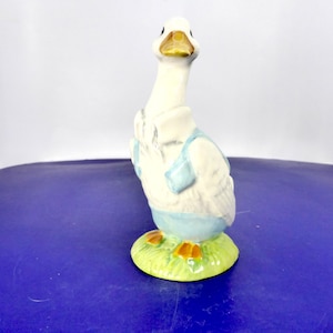 Puede incluir: Una figura de cerámica de un ganso blanco de pie. El ganso tiene un pico amarillo y lleva un chaleco azul claro y un cuello blanco. Está colocado sobre una base circular pintada con hierba verde y tiene patas de color naranja.