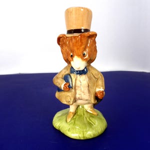 Puede incluir: Una figura de cerámica de un ratón antropomórfico que lleva un sombrero de copa beige, una chaqueta marrón claro, un chaleco blanco y una pajarita azul. El ratón está sobre una base verde y sostiene un pequeño libro azul con detalles pintados.