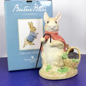 May include: A ceramic figurine of a rabbit wearing a red cape and holding a dark brown shepherd's crook, standing next to a woven basket filled with dark berries. The box behind it is light blue with the text Beatrix Potter Classics.
