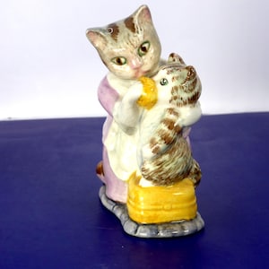 May include: A ceramic figurine depicting an adult cat with grey and brown tabby markings wearing a light purple garment, holding a smaller brown and white tabby kitten that is standing on a yellow rectangular base with a smooth glossy finish.