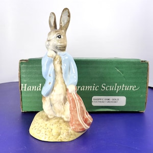 Peut inclure: Une figurine de lapin en céramique portant une veste bleu clair et un sac rouge, posée sur une base texturée. Derrière se trouve une boîte verte avec le texte Hand Painted Ceramic Sculpture et une étiquette RABPFC 5190 - GOLD PETER POCKET HIKERCHIEF.