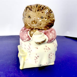 Peut inclure: Une figurine de hérisson en céramique avec un visage et des piquants bruns texturés, portant un gilet rose sur une robe crème à petits motifs floraux rouges. Le hérisson est assis sur un tabouret jaune et tient une petite tasse blanche.