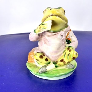 May include: A ceramic figurine of a seated frog wearing a light pink jacket and yellow trousers with brown spots. The frog has a yellow head, green markings, and is holding a small white object near its mouth while sitting on a green base.