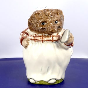Peut inclure: Une figurine en céramique représentant un hérisson brun portant un tablier blanc sur une chemise à carreaux bruns et blancs. Le hérisson porte une coiffe blanche à motifs floraux bleus et tient une petite cuillère argentée dans sa main gauche.