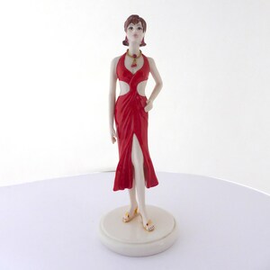 May include: A figurine of a woman in a red dress with a high slit and cut-out details. The figurine has a gold necklace and matching sandals. The figure is standing on a white circular base.