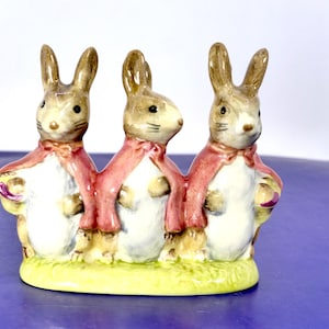 May include: A ceramic figurine featuring three brown rabbits standing side-by-side on a green base. Each rabbit has a white chest and wears a pink cape tied at the neck. The rabbits have pointed ears, dark eyes, and whiskers, all finished with a glossy glaze.