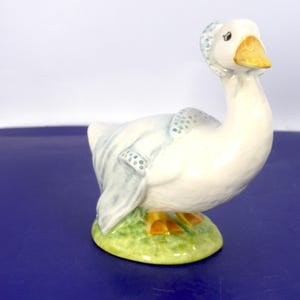 May include: A ceramic figurine of a white goose wearing a blue bonnet and a matching blue shawl with a dotted pattern. The goose has an orange beak and orange feet, and it stands on a textured green base that resembles a patch of grass.
