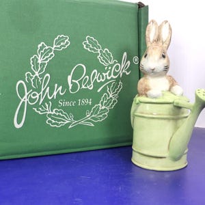 May include: A ceramic figurine of a brown and white rabbit sitting inside a light green watering can. The figurine is positioned next to a green cardboard box with the white text John Beswick Since 1894 and an oak leaf wreath logo.