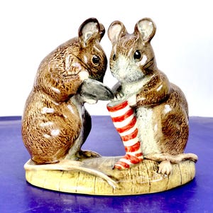May include: A ceramic figurine featuring two brown mice standing on a textured base. One mouse holds a dark gray object while the other holds a red and white striped stocking. The mice have light brown bellies, dark brown fur, and glossy finishes.