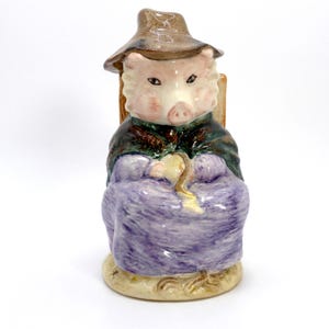 May include: A ceramic pig figurine wearing a hat and seated in a chair. The pig is dressed in a dark green shawl and a purple dress. The figurine has a light brown hat and a light brown base.