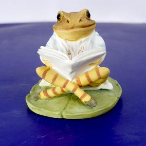 May include: A ceramic frog figurine sitting on a green lily pad. The frog has a tan head, wears a light blue jacket with pink floral details, and has yellow and brown striped legs. It is holding an open white book in its hands while sitting.