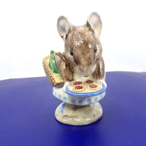 May include: A ceramic figurine of a brown mouse standing on a pedestal base. The mouse wears a white apron with blue polka dots and holds a blue tray containing three small round biscuits with red centres. A woven basket is visible on the mouse's back.