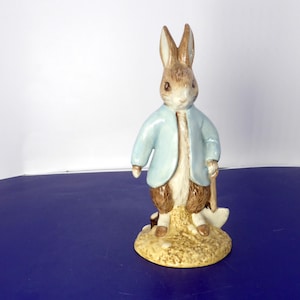 May include: A ceramic figurine of a brown rabbit standing on a textured tan base. The rabbit wears a light blue jacket and holds a small white shovel in one hand. The figure features detailed fur markings and painted facial features in brown and white.