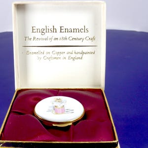 May include: A round enamel box with a gold-tone metal rim, featuring a hand-painted illustration of a pink spool of thread and sewing needles on a white background. It is presented in a white box with the text English Enamels, The Revival of an 18th Century Craft, Enamelled on Copper and handpainted by Craftsmen in England.
