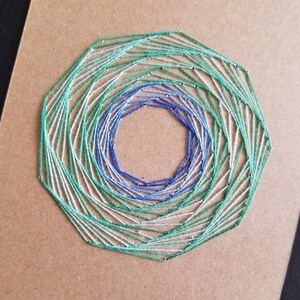 May include: A green, blue, and white string art design on a brown background. The design is a spiral pattern with a circular shape in the center.