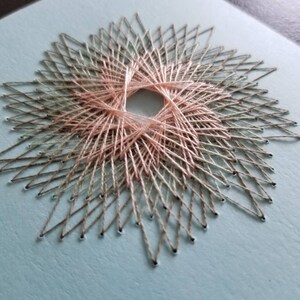 May include: A close-up of a string art design with a circular pattern in the center. The string is a light pink and a light green color. The background is a light blue.