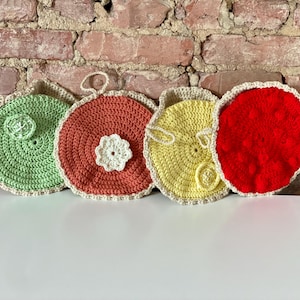 Könnte beinhalten: Vier gehäkelte Topflappen in verschiedenen Farben und Mustern. Die Topflappen sind grün, orange, gelb und rot. Jeder Topflappen hat eine weiße gehäkelte Blume in der Mitte.
