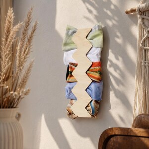 Puede incluir: Un organizador de pared de madera de color claro con forma de zigzag vertical, que cuenta con cinco ranuras que sostienen diademas de tela plegadas en varios colores y patrones, incluyendo verde claro, blanco, naranja, azul y un estampado de mármol marrón.