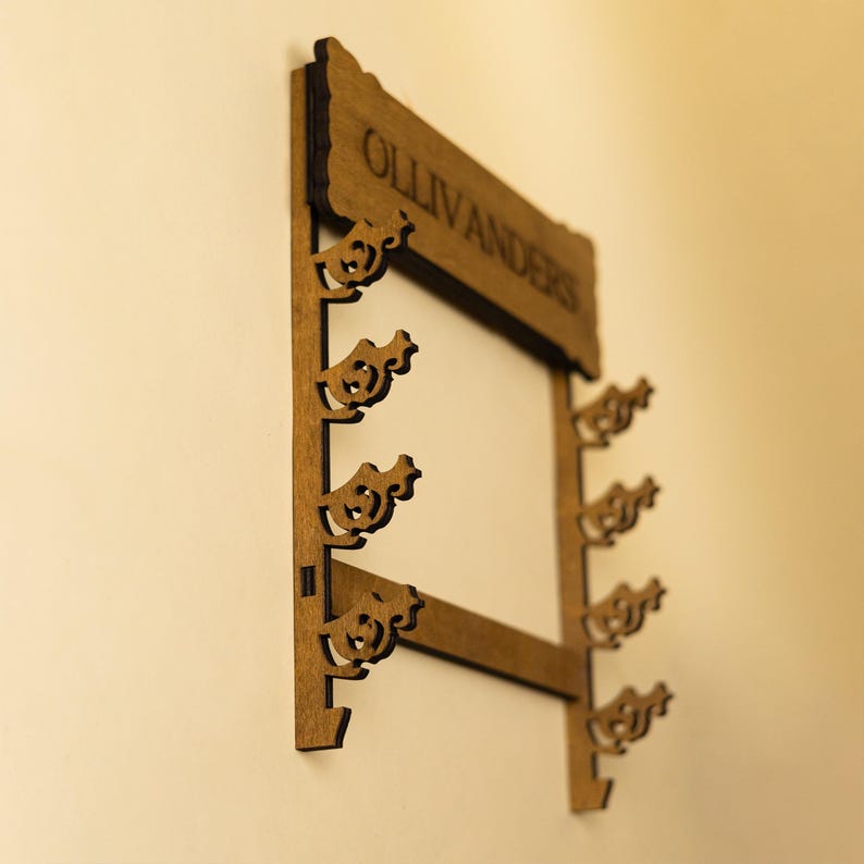 Puede incluir: Soporte de pared de madera para varitas con la palabra "Ollivanders" en la parte superior. El soporte tiene seis ranuras para varitas, cada una con forma de p&aacute;jaro estilizado.