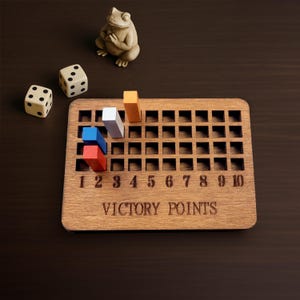 May include: A rectangular wooden board with a grid of square cutouts, labeled VICTORY POINTS and numbered 1 through 10. Three colored rectangular pegs in red, blue, and white are inserted into the grid, with an orange peg nearby. Two dice and a small frog figurine are present.